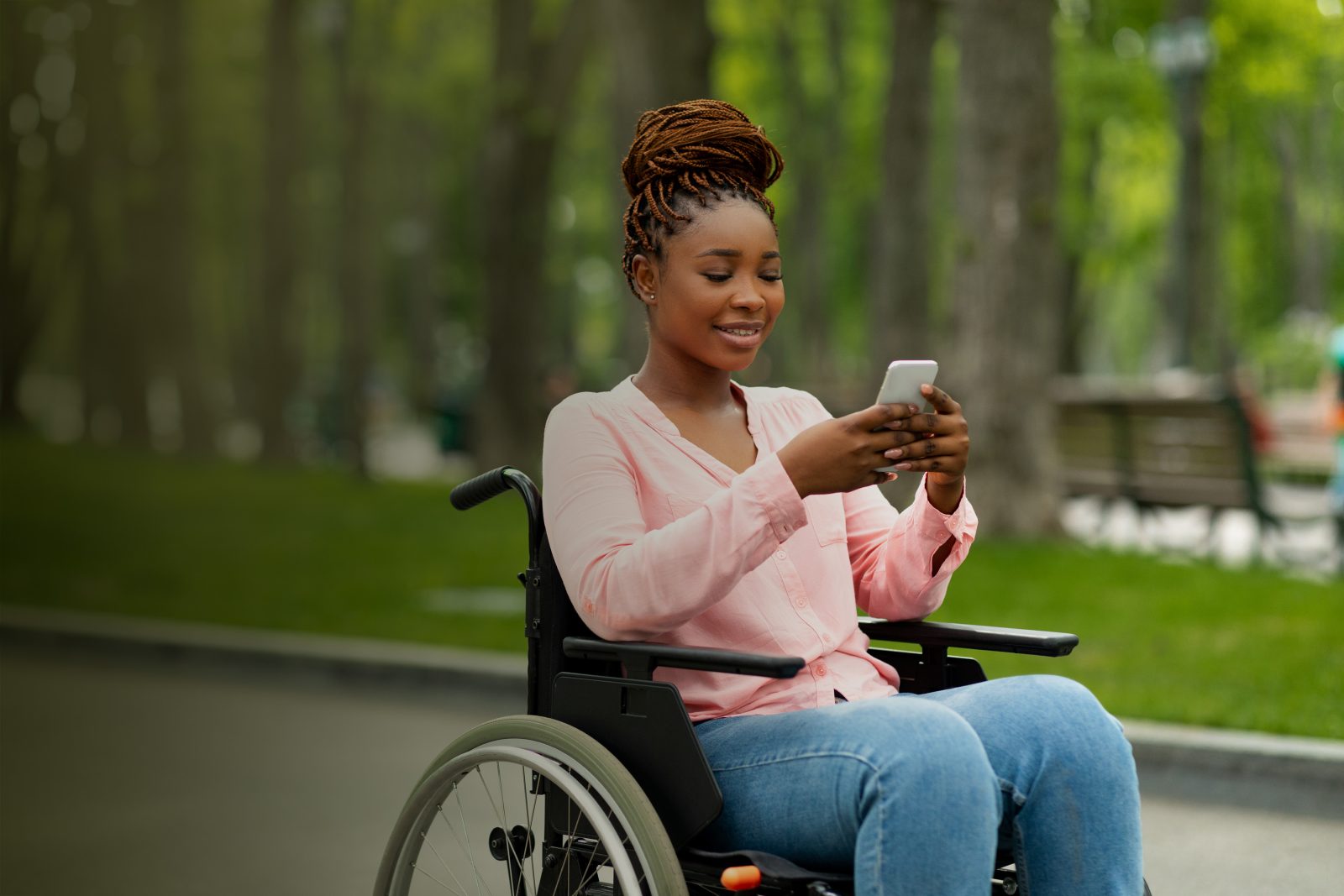Jeune fille en fauteuil roulant sur son téléphone cellulaire à regarder ses messages dans un parc de la ville, en souriant. 