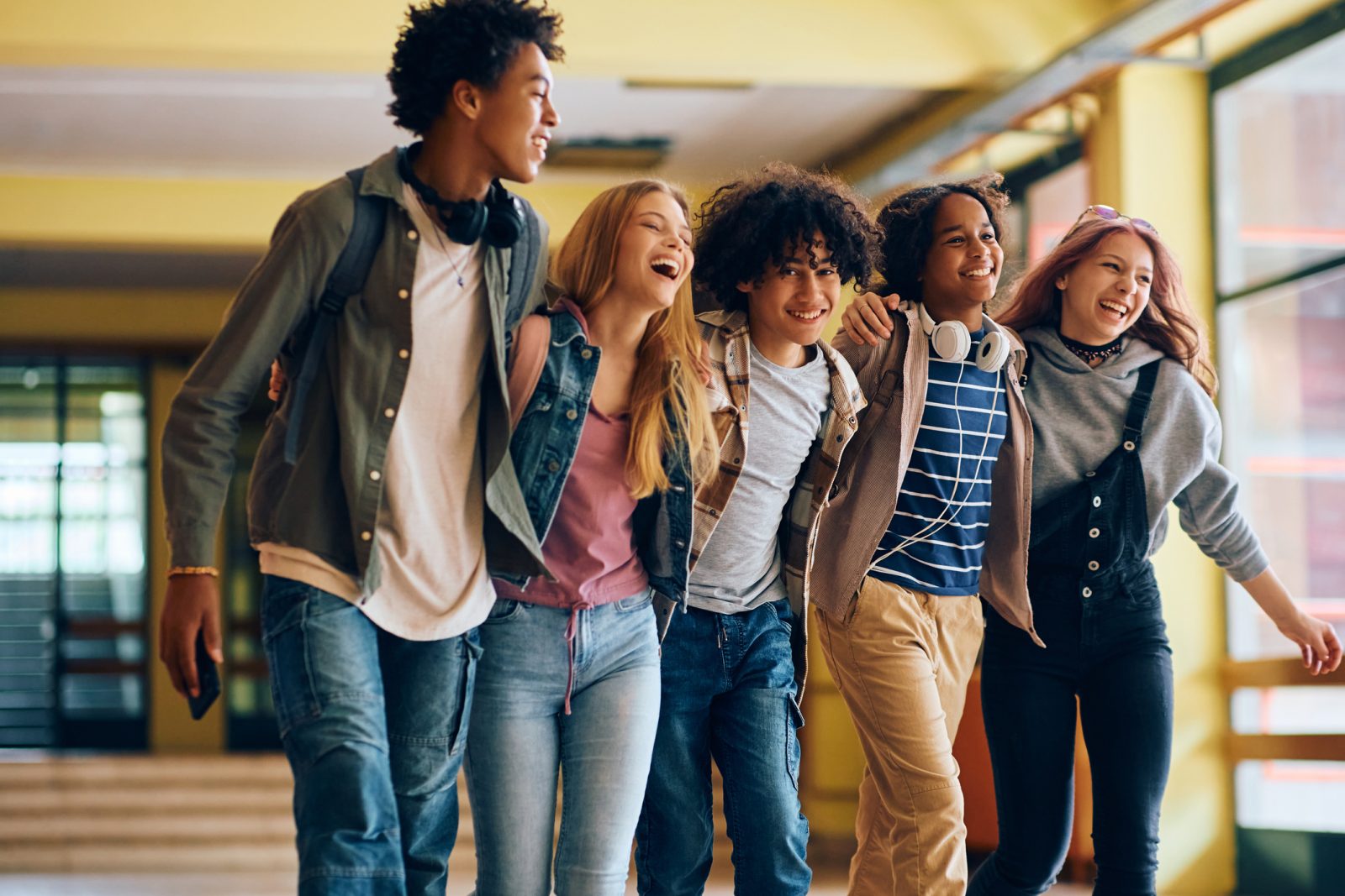 Groupe d’élèves de différentes origines se tenant par la taille et marchant joyeusement dans le corridor de l’école avec divers appareils numériques 