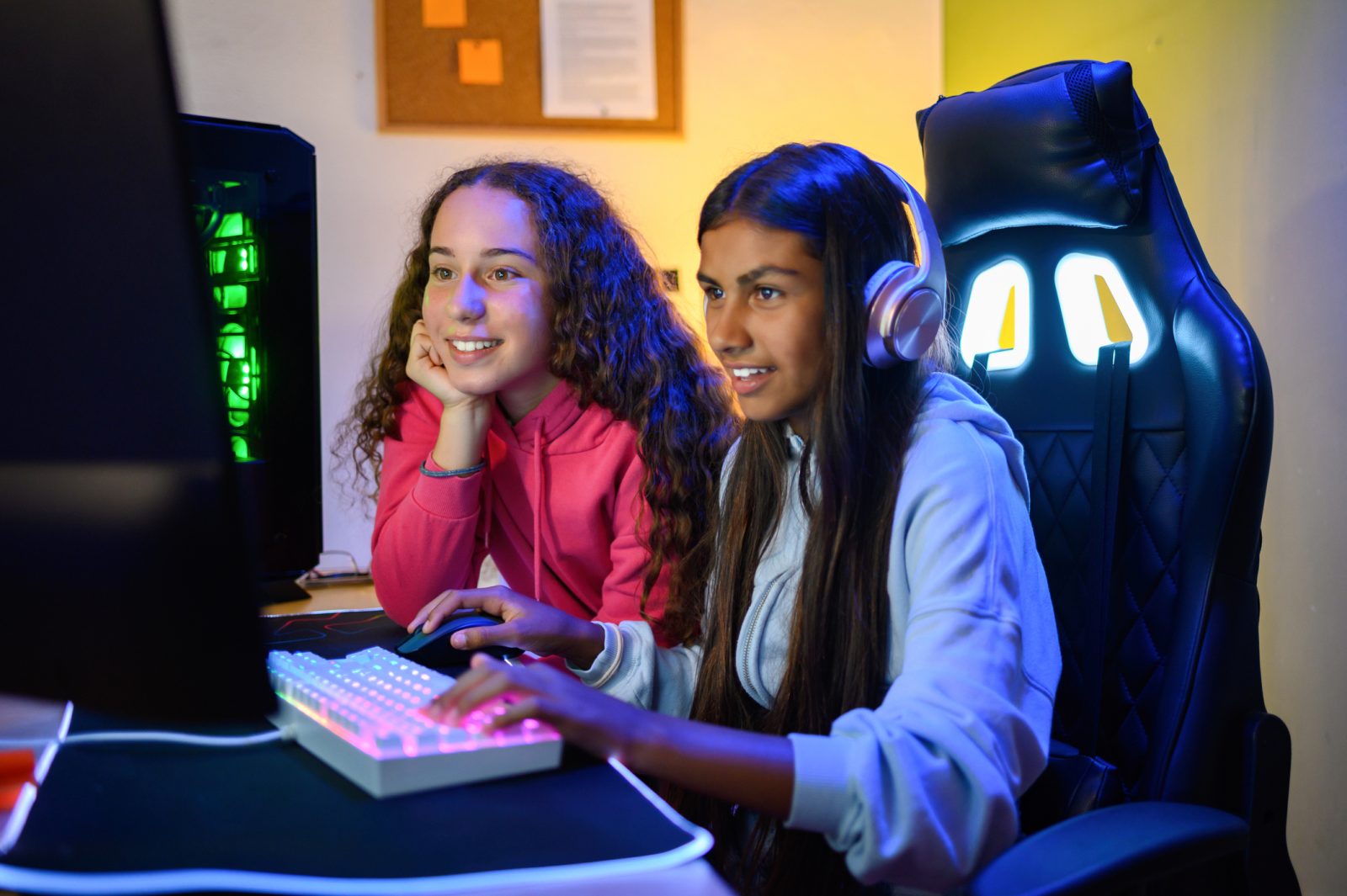 Positive teenage multiracial girls looking at screen of monitor while sitting at table with gaming PC and using keyboard to play game in room.